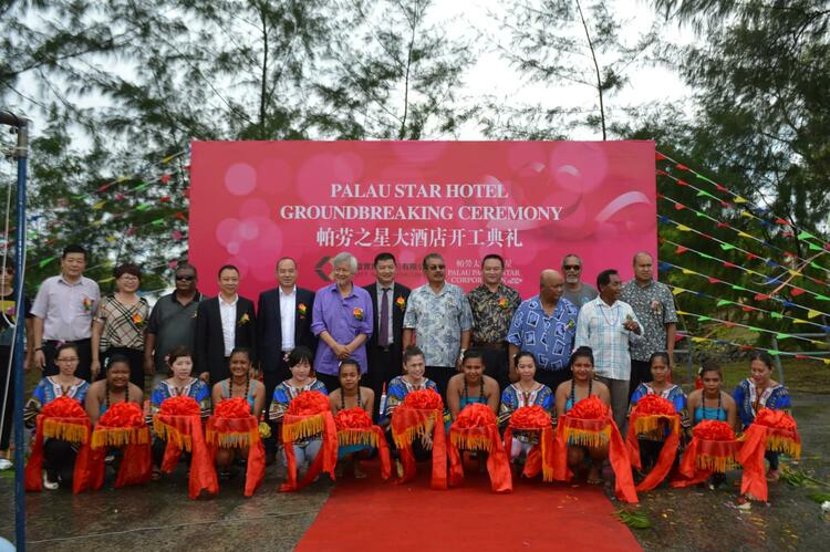 A line of people holding an orange ribon outside in front of a large pink sign for Palau Star Hotel Groundbreaking Ceremony