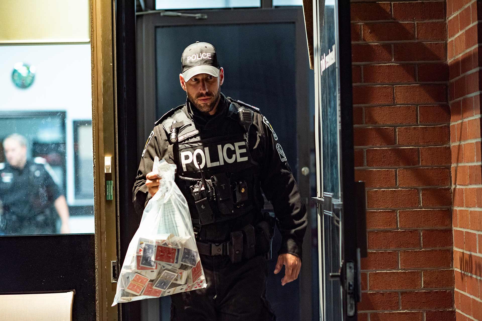 An officer emerges from a building holding alleged gambling paraphernalia qhiddkiqeiqqdinv